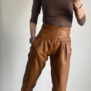 May include: A woman wearing a brown long-sleeve shirt and high-waisted, pleated brown leather pants. The pants have a wide waistband. She is wearing black and leopard print sneakers.