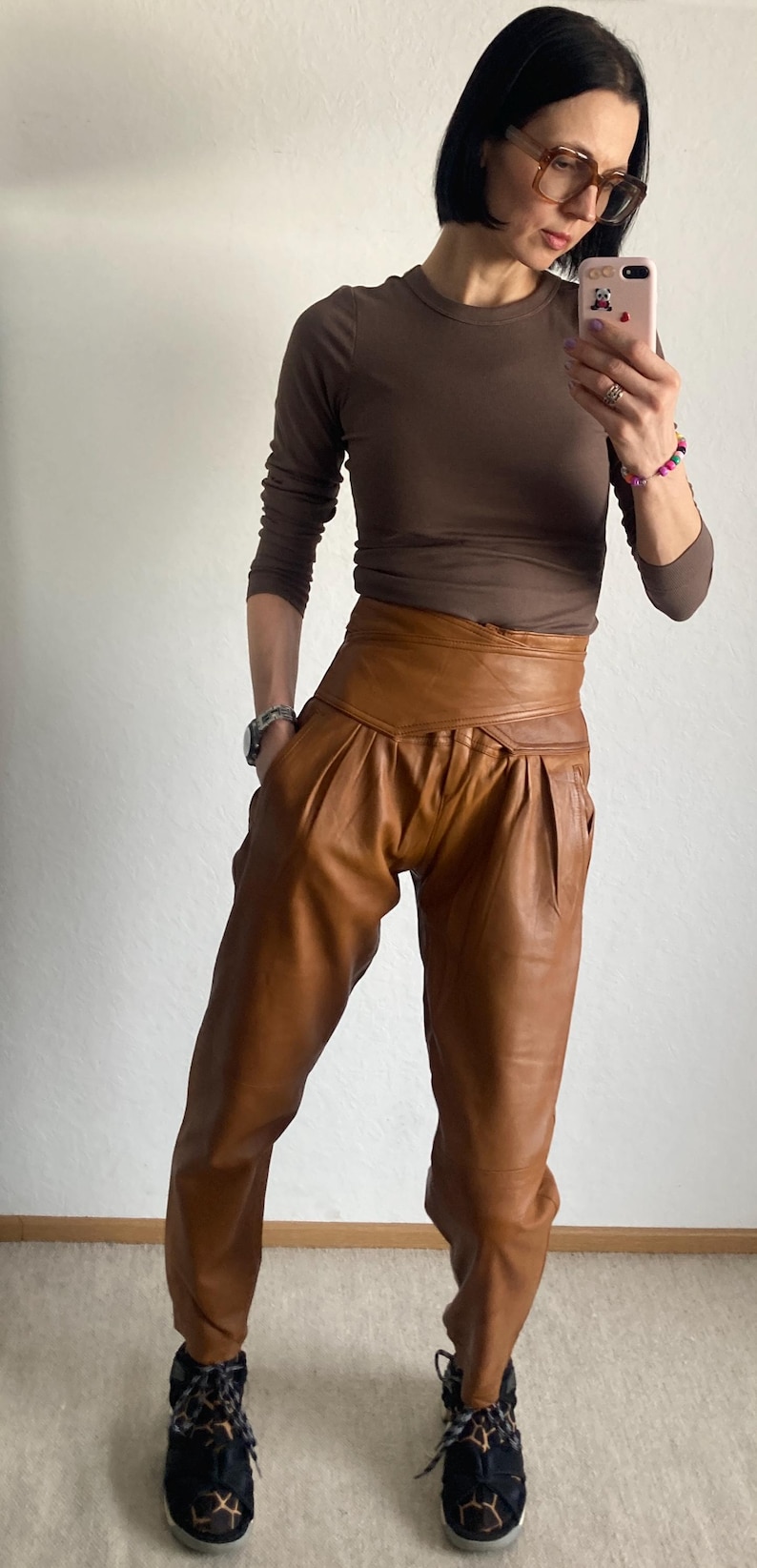 May include: A woman wearing a brown long-sleeve shirt and high-waisted, pleated brown leather pants. The pants have a wide waistband. She is wearing black and leopard print sneakers.