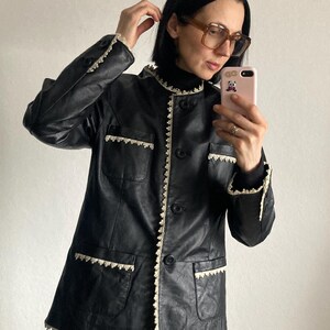 May include: Black leather jacket with a round collar and button closure. The jacket features cream-colored lace trim along the collar, pockets, and cuffs. The jacket has two front pockets.