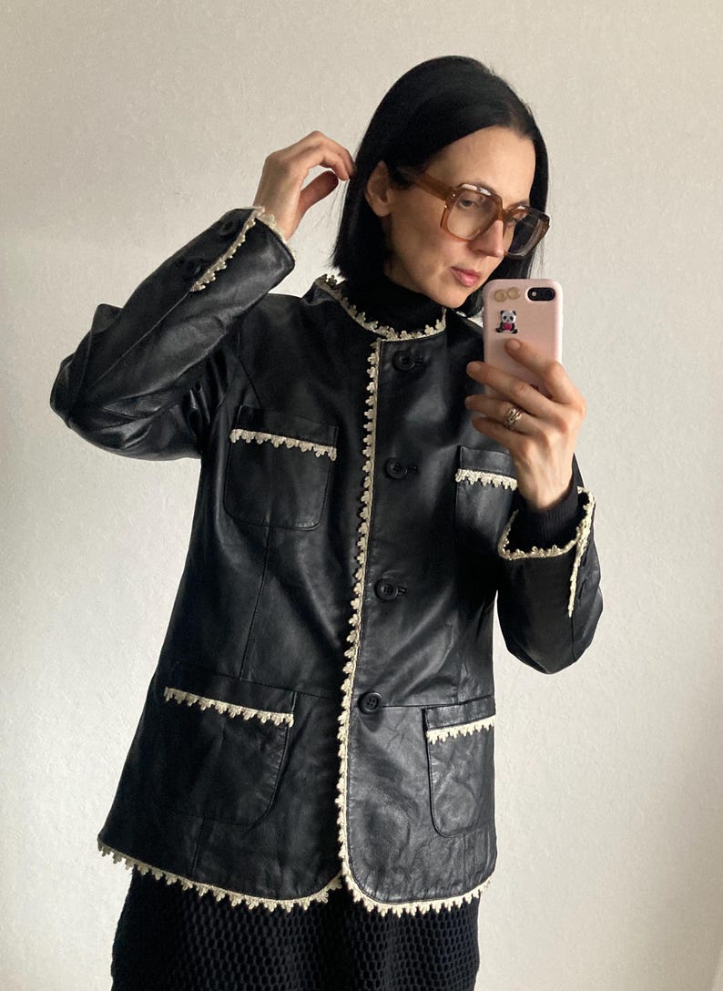 May include: Black leather jacket with a round collar and button closure. The jacket features cream-colored lace trim along the collar, pockets, and cuffs. The jacket has two front pockets.