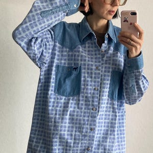 May include: A blue and white plaid button-up shirt with denim accents on the shoulders and pockets. The shirt features a small embroidered camel on the pocket. The long-sleeved shirt has a classic collar and buttoned cuffs.