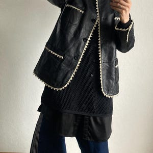May include: Black leather jacket with white trim, worn over a black top and dark blue pants. The jacket has a classic design with a button-up front and pockets. The pants have white stripes down the side. The outfit is completed with yellow sneakers.