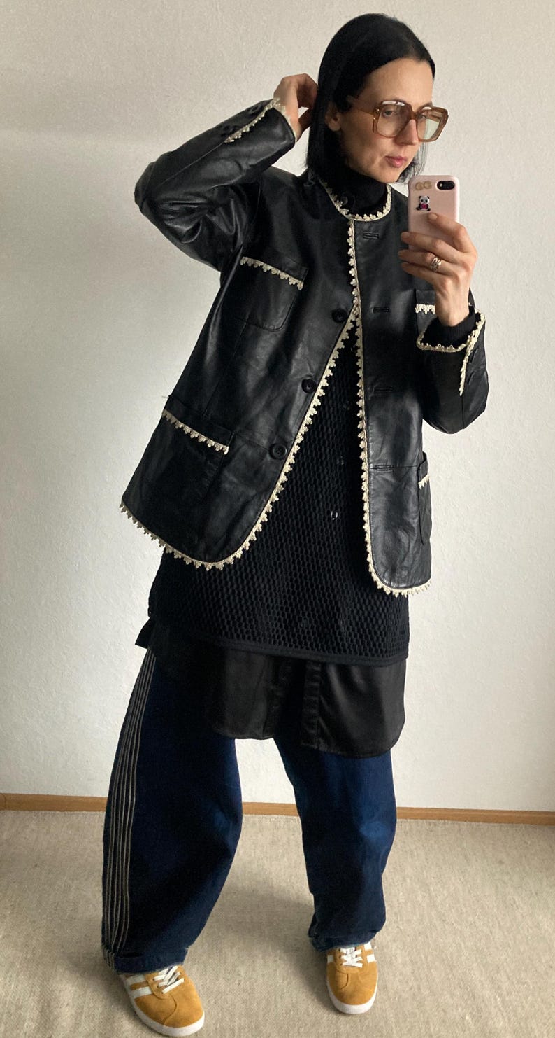 May include: Black leather jacket with white trim, worn over a black top and dark blue pants. The jacket has a classic design with a button-up front and pockets. The pants have white stripes down the side. The outfit is completed with yellow sneakers.