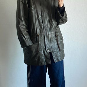 May include: A dark green leather jacket with a fur-trimmed hood. The jacket has a button-up front, pockets, and drawstring details. The person is wearing dark blue pants.