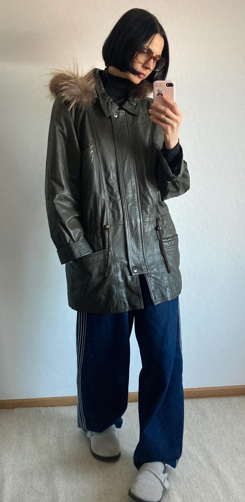 May include: A dark green leather jacket with a fur-trimmed hood. The jacket has a button-up front, pockets, and drawstring details. The person is wearing dark blue pants.