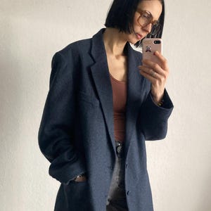 May include: A navy blue blazer with a traditional lapel and button fastening. The jacket has a loose fit and looks to be made from a wool mix. The model is wearing a brown top and grey trousers.