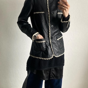 May include: Black leather jacket with cream-colored trim and pocket details. The jacket has a classic design with a button-up front. The model is wearing a black turtleneck, dark blue pants, and tan sneakers.