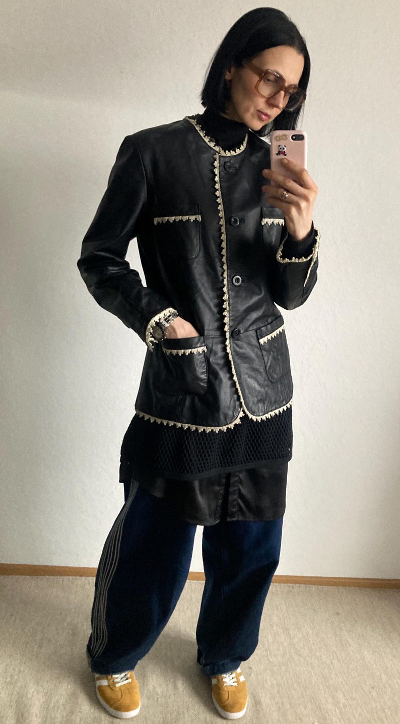 May include: Black leather jacket with cream-colored trim and pocket details. The jacket has a classic design with a button-up front. The model is wearing a black turtleneck, dark blue pants, and tan sneakers.