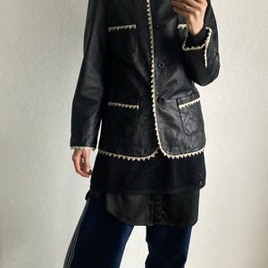 May include: Black leather jacket with white trim and a black turtleneck. The jacket has a classic design with a button closure and pockets. The person is wearing dark blue pants with white stripes and yellow sneakers.