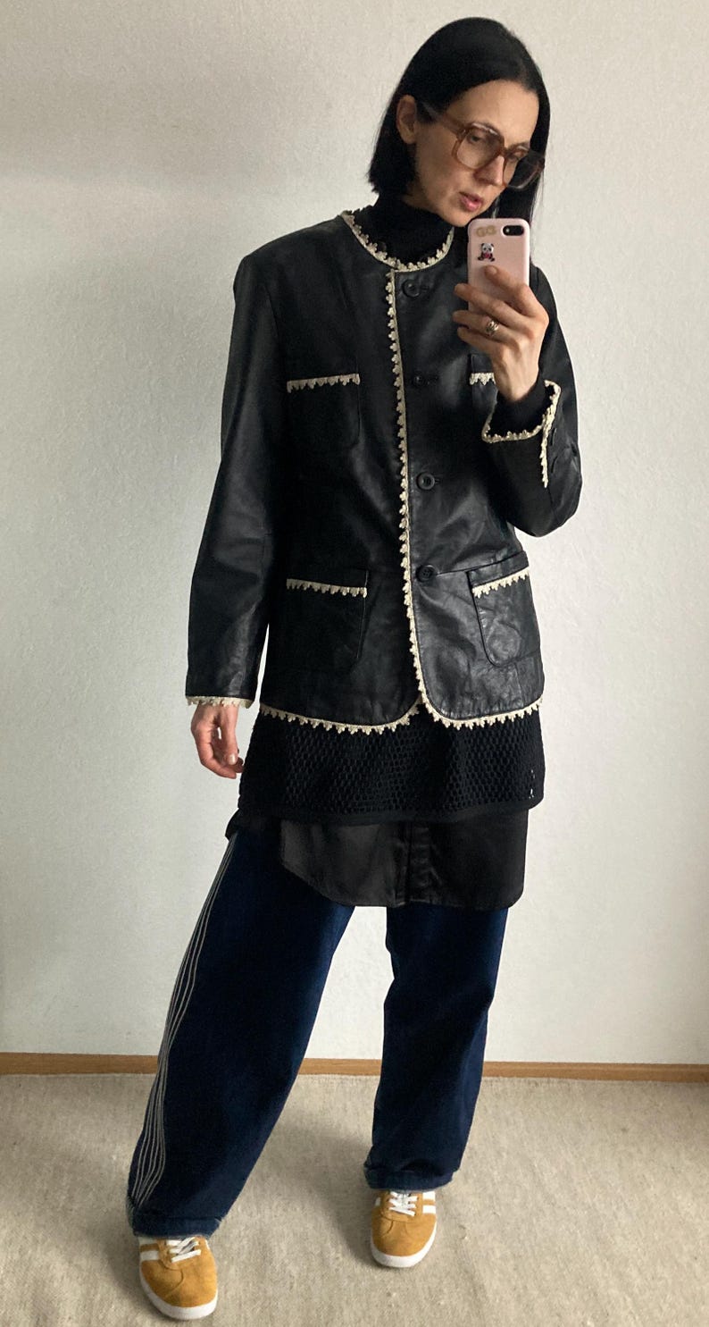 May include: Black leather jacket with white trim and a black turtleneck. The jacket has a classic design with a button closure and pockets. The person is wearing dark blue pants with white stripes and yellow sneakers.