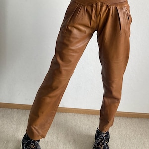 May include: Brown leather pants with a high waist and pleated front. The pants have a unique wrap-around design at the waist and pockets. The model is wearing black and brown patterned sneakers.