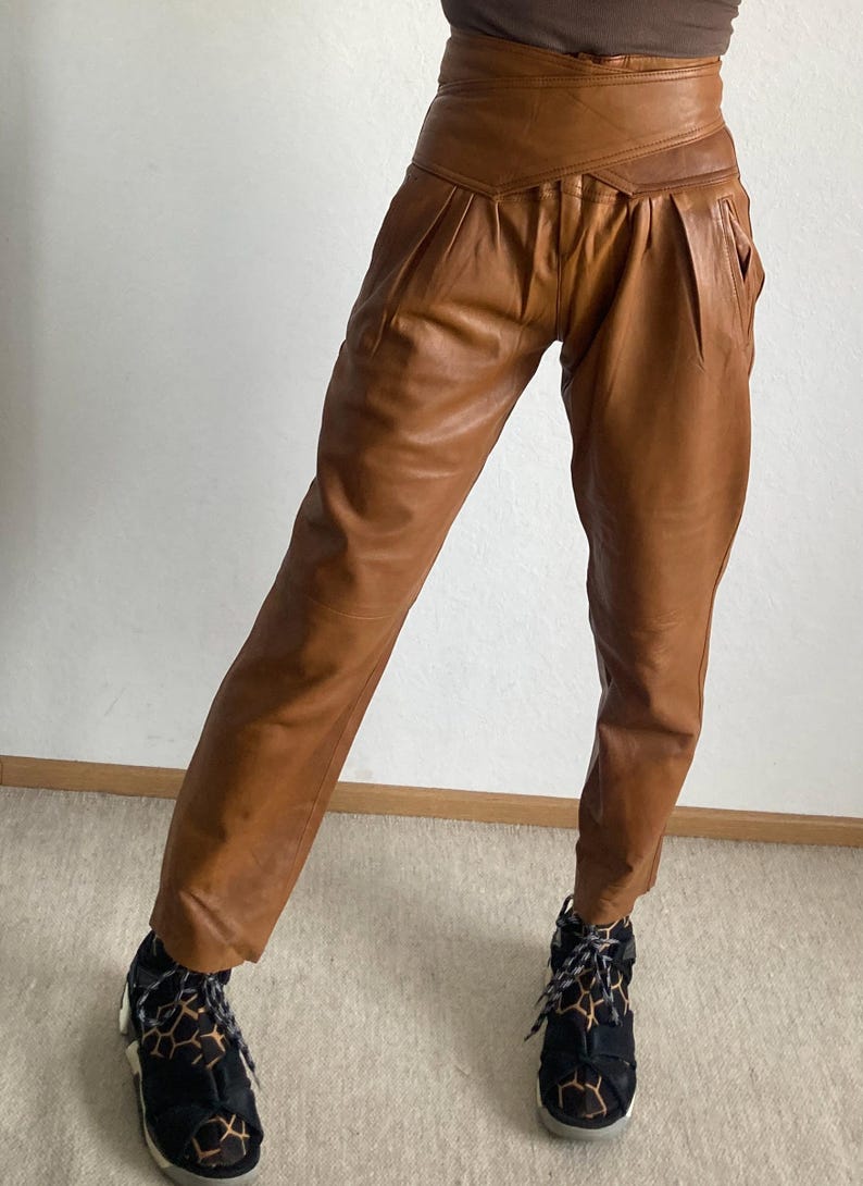 May include: Brown leather pants with a high waist and pleated front. The pants have a unique wrap-around design at the waist and pockets. The model is wearing black and brown patterned sneakers.