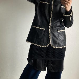 May include: Black leather jacket with white trim and pockets. The jacket is paired with a black top, dark blue pants, and yellow sneakers. The jacket has a button-up front and a collar.