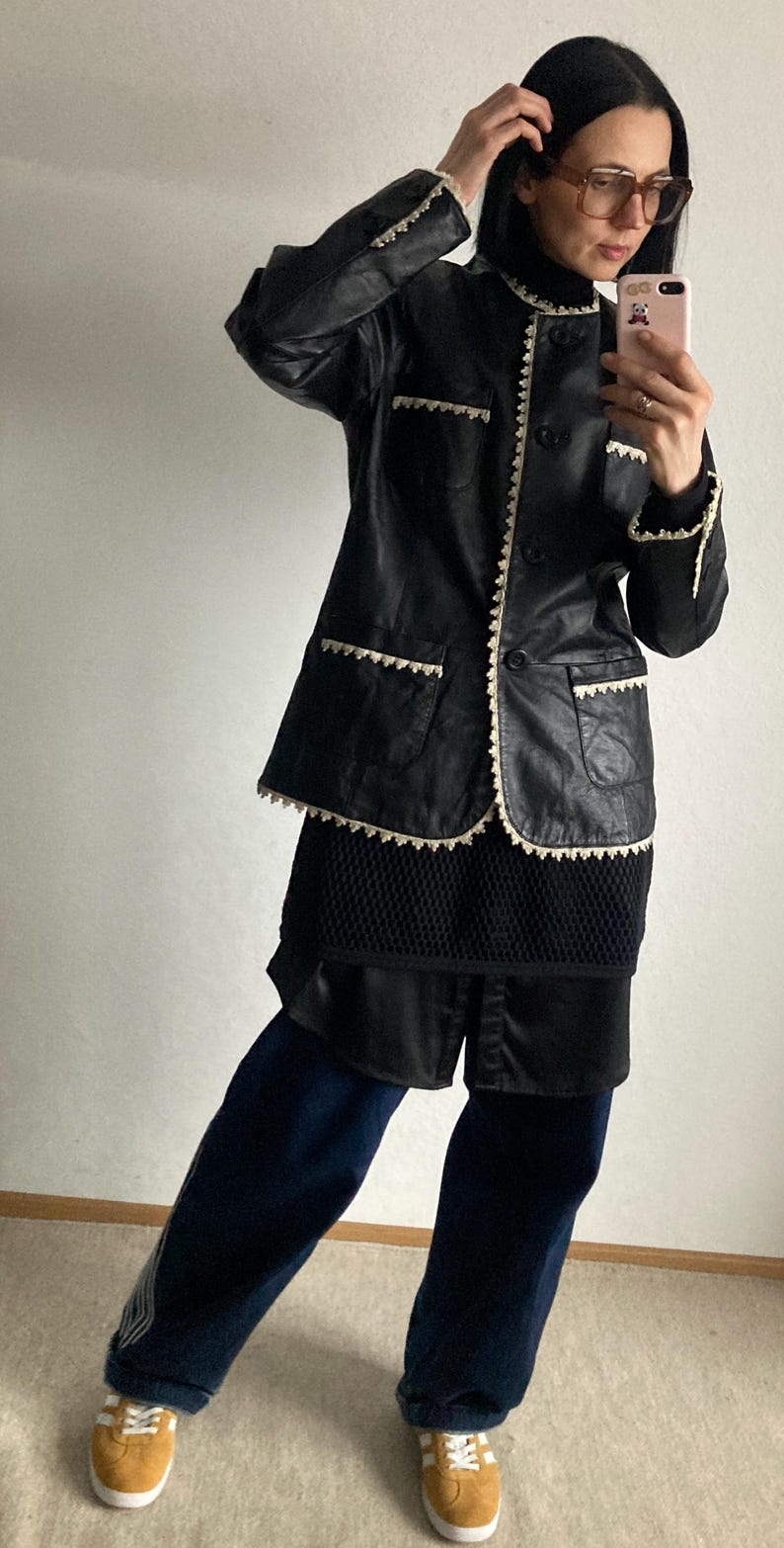 May include: Black leather jacket with white trim and pockets. The jacket is paired with a black top, dark blue pants, and yellow sneakers. The jacket has a button-up front and a collar.