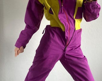 May include: A vintage ski suit in purple and yellow. The one-piece suit has a high collar, a zip-up front, and a fitted waist. The sleeves and legs are long, and the suit is worn with black boots. The suit is a retro style.