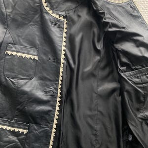 May include: Black leather jacket with cream-colored lace trim along the edges, pockets, and collar. The jacket has a satin lining and a button-up front. The jacket is open, revealing the interior lining and pockets.