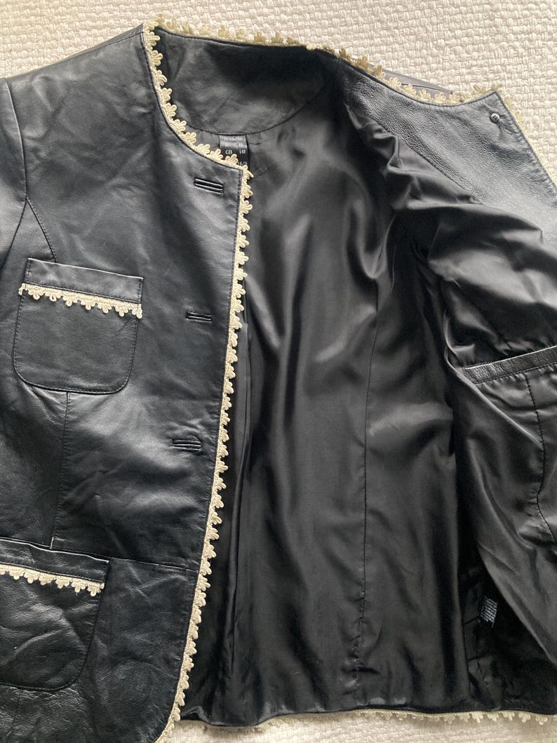 May include: Black leather jacket with cream-colored lace trim along the edges, pockets, and collar. The jacket has a satin lining and a button-up front. The jacket is open, revealing the interior lining and pockets.