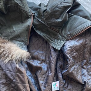 May include: Dark olive green leather jacket with a brown quilted lining. The jacket features a fur trim around the hood and a zipper closure. The interior lining is a dark brown color. A tag is visible on the inside.