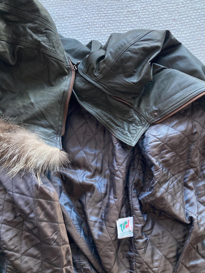 May include: Dark olive green leather jacket with a brown quilted lining. The jacket features a fur trim around the hood and a zipper closure. The interior lining is a dark brown color. A tag is visible on the inside.
