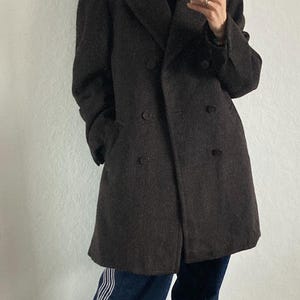 May include: A dark brown double-breasted wool coat. The coat is worn over a black turtleneck and paired with dark blue jeans with white stripes down the side. Red sneakers complete the look.