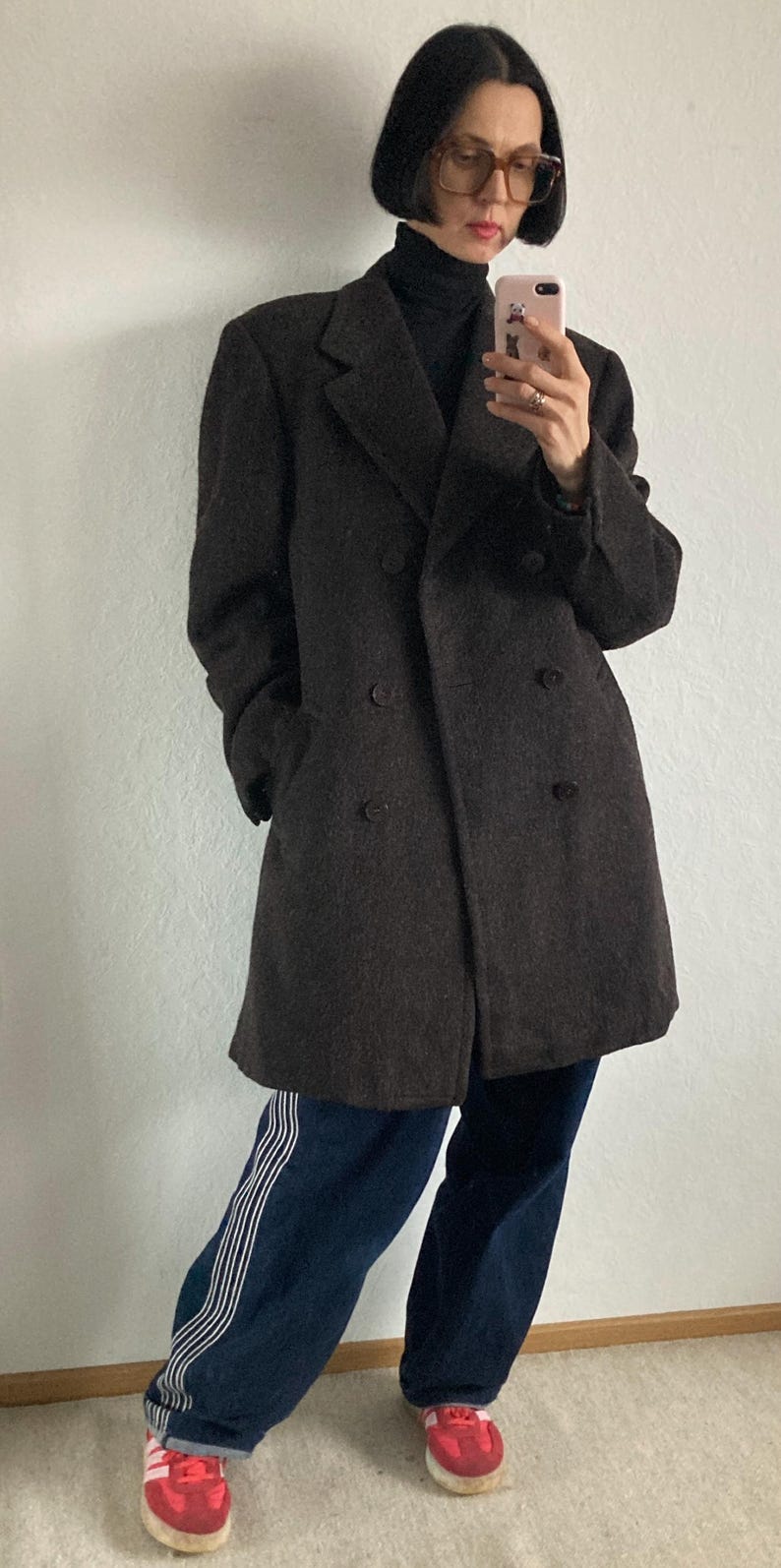May include: A dark brown double-breasted wool coat. The coat is worn over a black turtleneck and paired with dark blue jeans with white stripes down the side. Red sneakers complete the look.