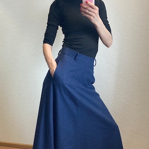 Op de afbeelding: Een vrouw draagt een zwarte turtleneck trui en een donkerblauwe maxi-rok met een naad aan de voorkant. Ze draagt zwarte schoenen en heeft haar haar naar achteren getrokken.