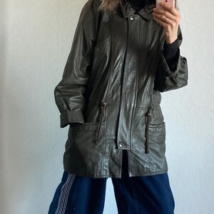 May include: A dark green leather jacket with a fur-lined hood. The jacket has a zipper closure, pockets, and drawstring details. The person is wearing dark blue denim pants with white stripes and gray suede slip-on shoes.