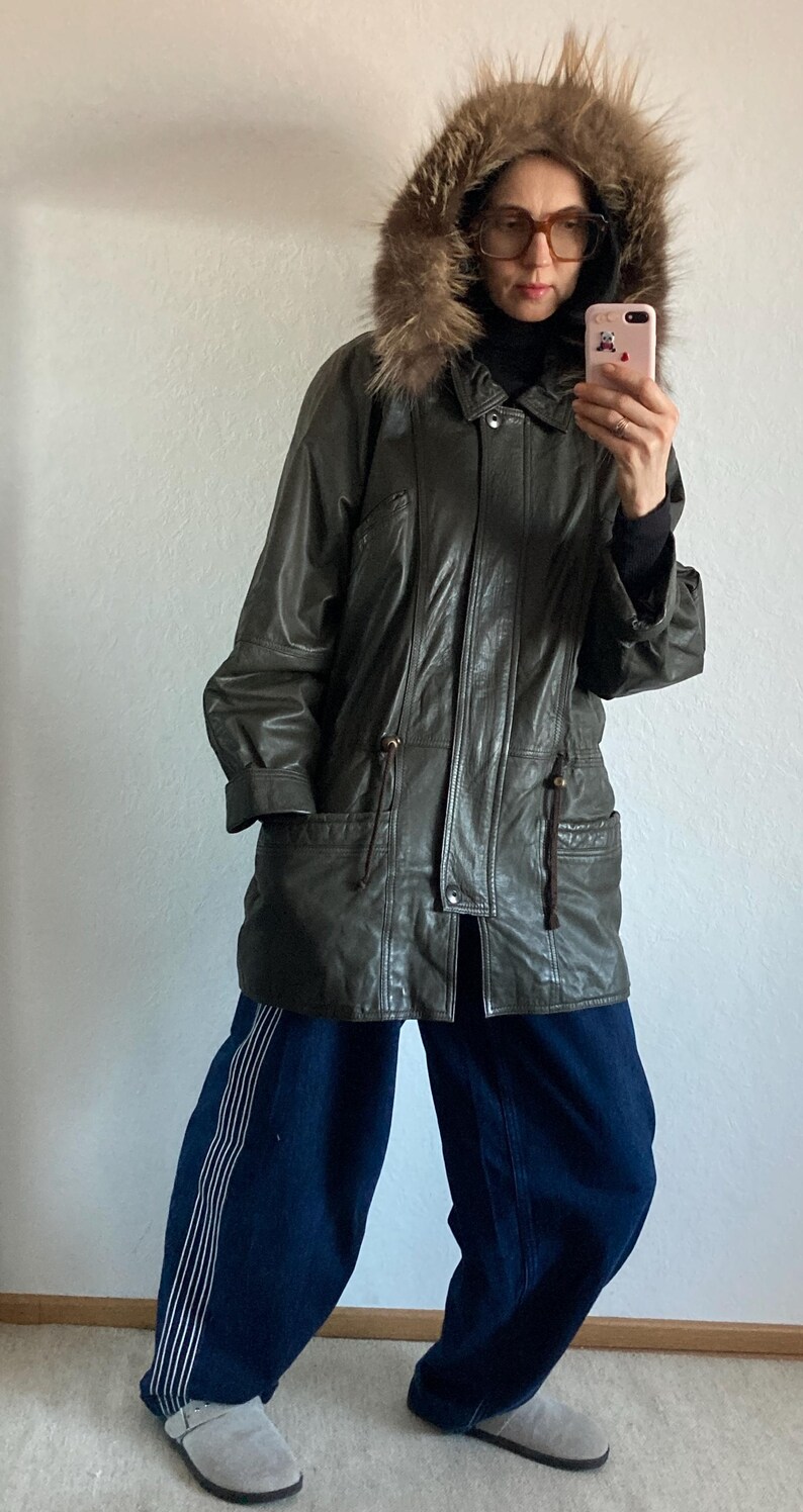 May include: A dark green leather jacket with a fur-lined hood. The jacket has a zipper closure, pockets, and drawstring details. The person is wearing dark blue denim pants with white stripes and gray suede slip-on shoes.