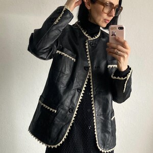 May include: Black leather jacket with cream-colored trim and button closure. The jacket has a round collar and pockets. The trim is a scalloped design. The person in the image is wearing a black turtleneck and glasses.