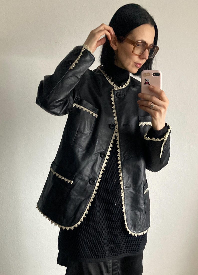 May include: Black leather jacket with cream-colored trim and button closure. The jacket has a round collar and pockets. The trim is a scalloped design. The person in the image is wearing a black turtleneck and glasses.
