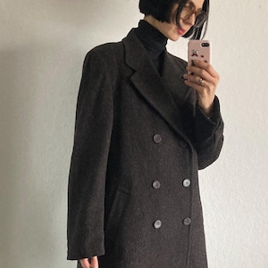 May include: A dark brown double-breasted coat with four buttons. The coat has a classic lapel and a tailored fit. The fabric appears to be a textured wool blend. The coat is worn over a black turtleneck.