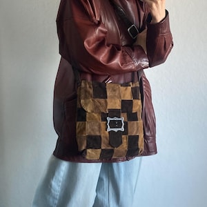 May include: A brown and tan checkered shoulder bag with a silver buckle. The bag is worn over a burgundy leather jacket and light blue jeans. The bag's design features a patchwork pattern.