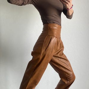 May include: A brown long-sleeve top and high-waisted, tan leather pants. The outfit is paired with black and tan patterned sneakers. The person is wearing glasses and a watch.