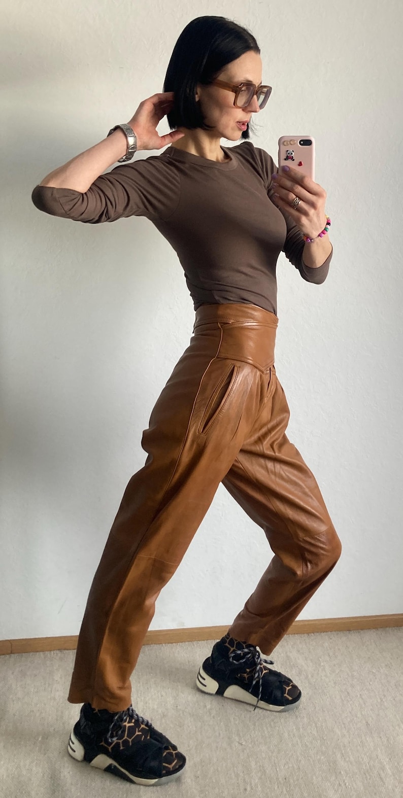 May include: A brown long-sleeve top and high-waisted, tan leather pants. The outfit is paired with black and tan patterned sneakers. The person is wearing glasses and a watch.