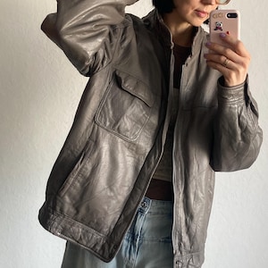 Op de afbeelding: Een grijze leren jas met twee voorzakken en een ritssluiting. De jas heeft een klassiek ontwerp en een licht versleten uiterlijk. De jas wordt gecombineerd met lichtblauwe jeans.