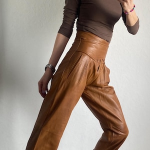 May include: A brown long-sleeve top and high-waisted, tan leather pants. The pants have a pleated design. The person is wearing black and white sneakers with a unique pattern. The outfit is a stylish ensemble.