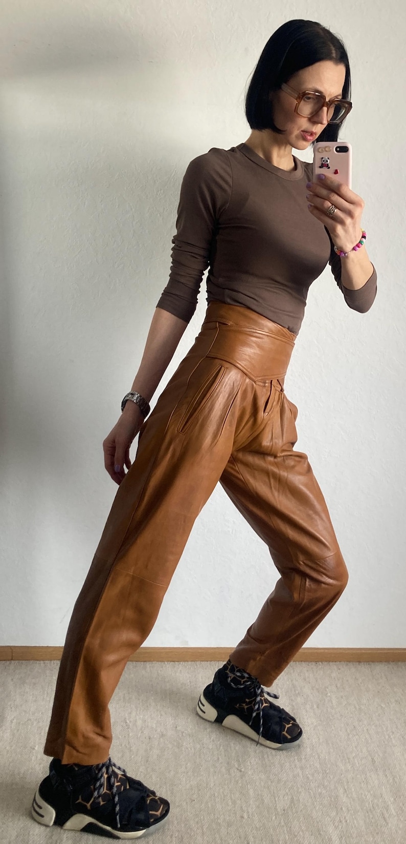 May include: A brown long-sleeve top and high-waisted, tan leather pants. The pants have a pleated design. The person is wearing black and white sneakers with a unique pattern. The outfit is a stylish ensemble.