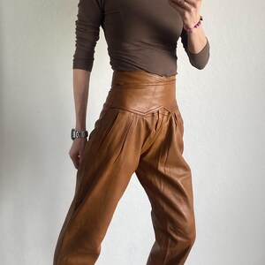 May include: A person wearing a brown long-sleeve shirt and high-waisted brown leather pants. The pants have a pleated design. The person is also wearing black and white sneakers with a patterned design.