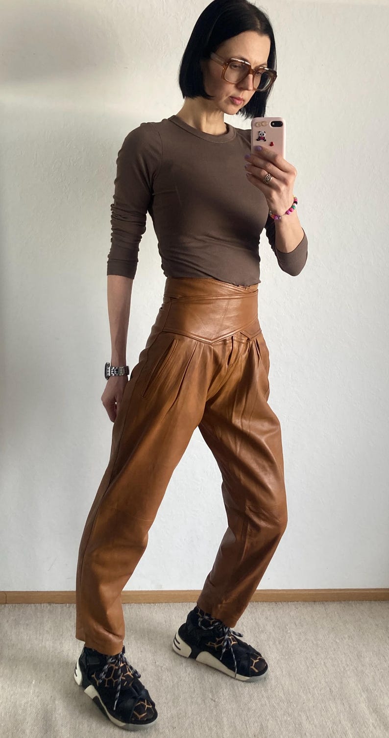 May include: A person wearing a brown long-sleeve shirt and high-waisted brown leather pants. The pants have a pleated design. The person is also wearing black and white sneakers with a patterned design.