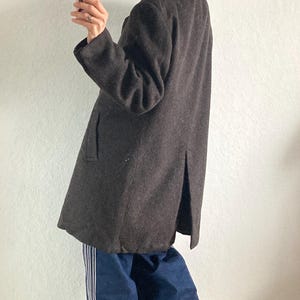 May include: A dark brown wool coat with a classic silhouette. The coat features a notched collar, long sleeves, and a knee-length hem. The person is wearing blue jeans with white stripes down the side.