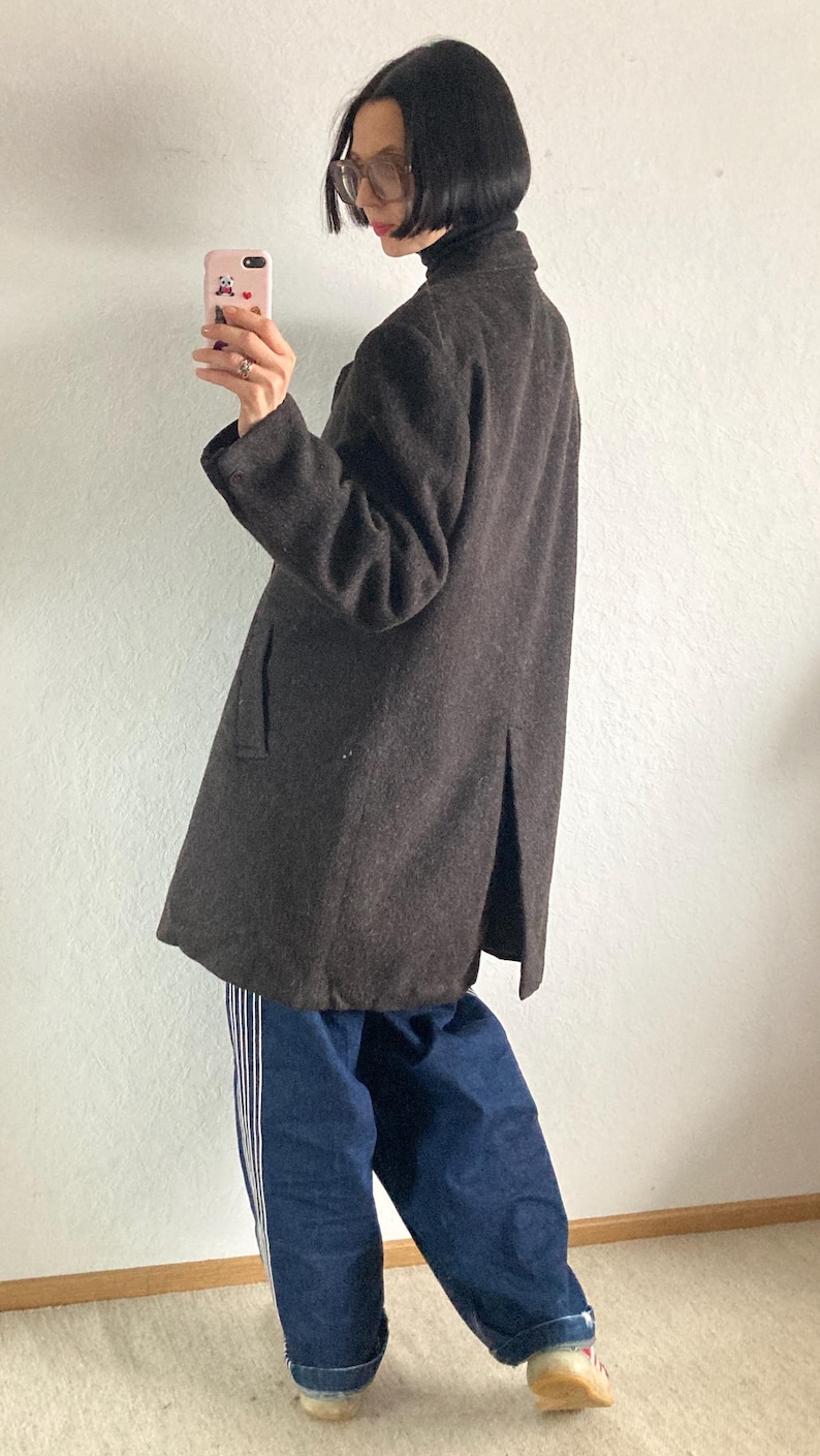 May include: A dark brown wool coat with a classic silhouette. The coat features a notched collar, long sleeves, and a knee-length hem. The person is wearing blue jeans with white stripes down the side.