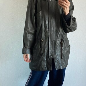 May include: A dark green leather jacket with a fur-trimmed hood. The jacket has a button closure, pockets, and a drawstring waist. The person in the image is wearing dark blue pants and gray shoes. The jacket is a vintage style.