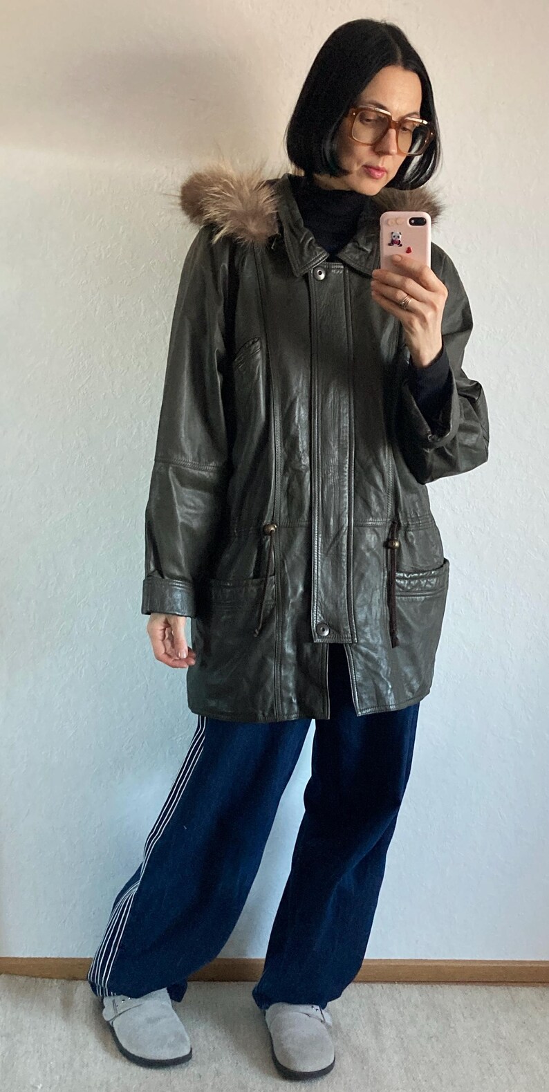 May include: A dark green leather jacket with a fur-trimmed hood. The jacket has a button closure, pockets, and a drawstring waist. The person in the image is wearing dark blue pants and gray shoes. The jacket is a vintage style.