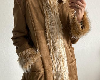 Könnte beinhalten: Ein langer, brauner Wildledermantel mit cremefarbenem Kunstfellbesatz am Kragen, an den Manschetten und vorne. Der Mantel hat ein klassisches Design mit Taschen und einer taillierten Passform, ideal für einen Vintage- oder Bohème-Look.