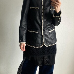 May include: Black leather jacket with cream-colored trim and pockets. The jacket is worn over a black top and paired with navy blue pants with white stripes. The outfit is completed with mustard-colored sneakers.