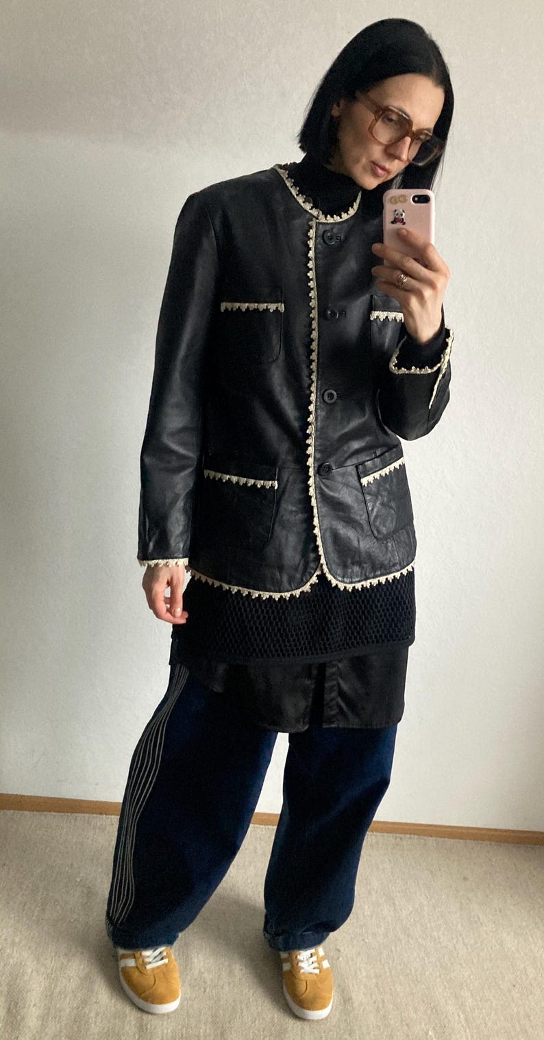May include: Black leather jacket with cream-colored trim and pockets. The jacket is worn over a black top and paired with navy blue pants with white stripes. The outfit is completed with mustard-colored sneakers.