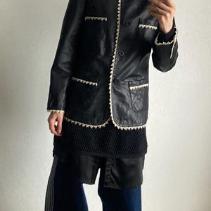 May include: Black leather jacket with cream-colored trim and pockets. The jacket has a round collar and button closure. The person is wearing a black top, dark blue pants with white stripes, and tan sneakers.