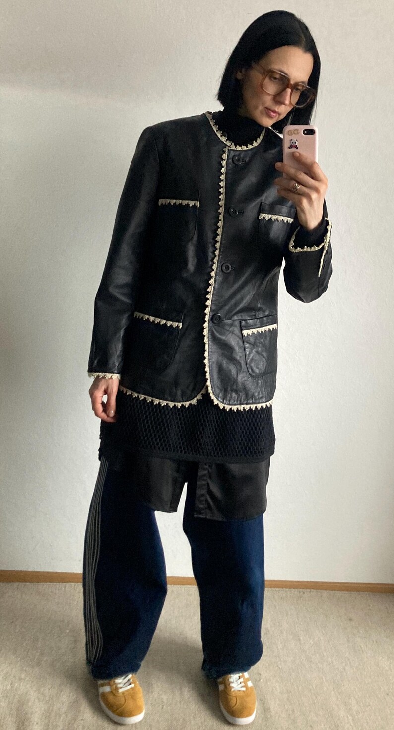 May include: Black leather jacket with cream-colored trim and pockets. The jacket has a round collar and button closure. The person is wearing a black top, dark blue pants with white stripes, and tan sneakers.