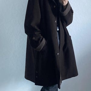 May include: A brown long-sleeved coat with a button closure and a hood. The coat is worn over a black turtleneck and blue jeans. The person is wearing white and brown fuzzy slippers.