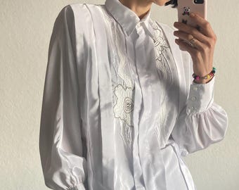 May include: White button-down shirt with intricate floral embroidery on the front. The long-sleeved blouse features pleated details and a classic collar. The cuffs are buttoned and the sleeves are puffy.